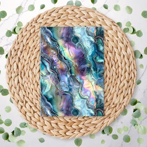 May include: An art print with an abstract design in shades of blue, teal, purple, and gold. The print is placed on a woven, circular placemat. The design has swirling patterns. Green leaves are scattered around.