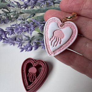 May include: A heart-shaped earring and a matching cookie cutter. The earring is white with a pink jellyfish design and a gold-colored clasp. The cookie cutter is pink and also features a jellyfish design. The background includes lavender.