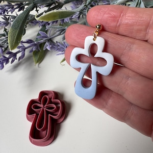 May include: A handmade, cross-shaped earring with a gradient of white to light blue, held by a hand. A matching cross-shaped clay cutter is next to the earring. The earring has a gold-colored stud.