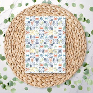 May include: A notebook with a marine-themed pattern featuring crabs, fish, and other sea elements in blue, orange, and yellow. The notebook rests on a woven, circular placemat, surrounded by green leaves. The notebook is approximately 20 cm x 15 cm.