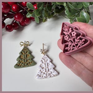 May include: Christmas-themed earrings and a cookie cutter. One earring is olive green with gold accents, the other is white with gold flecks. A pink tree-shaped cookie cutter with a floral design is held in a hand. Red berries and green leaves are in the background.
