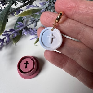 May include: A gold hoop earring with a round, white and blue pendant featuring a raised cross design. A pink, circular object with a cross-shaped indentation sits below the earring. The earring is held by a hand.