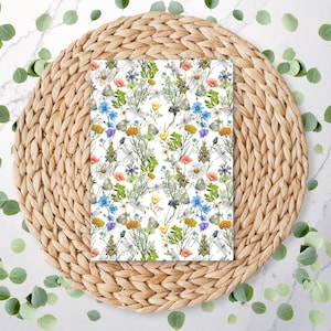 May include: A white background with a floral pattern featuring various flowers and leaves in shades of blue, yellow, pink, and green. The pattern is displayed on a woven, round, light brown coaster.