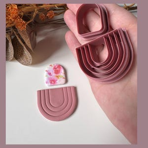 May include: Pink polymer clay cutters in a rainbow shape, with a floral patterned clay piece. The cutters are held in a hand, with a dried flower arrangement in the background. The cutters are for making earrings or other jewellery.