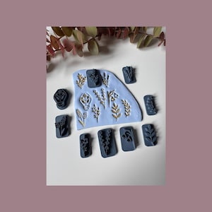 May include: A selection of dark blue clay stamps featuring botanical designs, such as leaves and flowers, displayed on a light blue clay base. Gold-coloured botanical designs are also visible. The background is white with a mauve border.