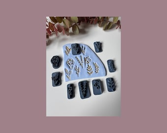 May include: A collection of dark blue clay stamps with botanical designs, including leaves and flowers, arranged on a light blue clay base. Gold-colored botanical designs are also present. The background is white with a mauve border.