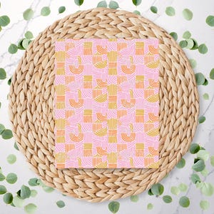 May include: A pink notebook featuring a geometric pattern of yellow and orange arches and lines. The notebook sits on a woven, circular placemat, with green leaves scattered on a white marble surface.