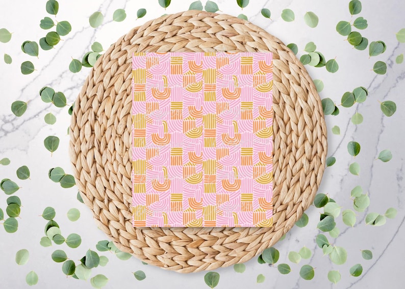 Może przedstawiać: R&oacute;żowy notatnik z geometrycznym wzorem ż&oacute;łtych i pomarańczowych łuk&oacute;w i linii. Notatnik znajduje się na plecionym, okrągłym podkładzie, otoczony zielonymi liśćmi na białej marmurowej powierzchni.