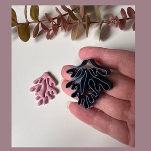 May include: Two abstract leaf-shaped brooches. One is a light pink colour, and the other is dark blue with white outlines. The brooches are on a white surface with a sprig of greenery in the background.