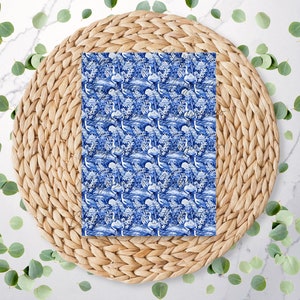 May include: A rectangular fabric with a blue and white toile pattern featuring birds, trees, and water elements. The fabric is placed on a round, woven, natural-coloured placemat, surrounded by scattered green eucalyptus leaves.