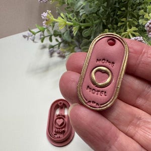 May include: A pink, oval-shaped cookie cutter with the word "HOME" and "MOTEL" embossed in a serif font. The cutter has a gold-colored border and a heart-shaped cutout. A second, smaller cutter is also visible.