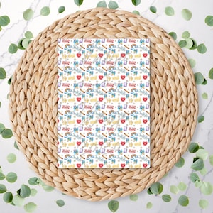May include: A white paper featuring a repeating pattern of rainbows, hearts, crowns, and text. The paper rests on a woven, circular placemat. Green leaves are scattered around the placemat.