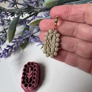 Peut inclure: Boucle d'oreille en argile beige avec fermoir doré, ornée d'un motif de feuille et d'une bordure de petites perles rondes. Un moule en argile rose assorti est également visible. La boucle d'oreille est tenue dans une main, avec des brins de lavande en arrière-plan.