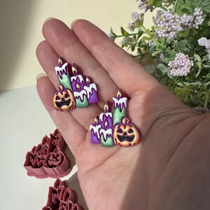 May include: A pair of earrings featuring a sculpted design of three candles with melting white wax and a carved jack-o'-lantern, colored in purple, green, and orange. Below, two matching pink plastic cookie cutters are visible on a flat surface.
