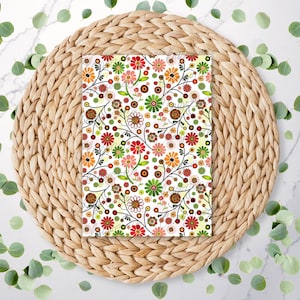 May include: A white background with a woven wicker coaster. On top of the coaster is a square piece of paper with a repeating pattern of red, orange, yellow, green, and brown flowers on white stems. The flowers are stylised and have a retro look.
