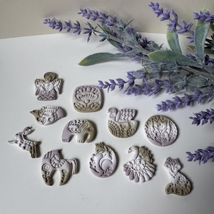 Peut inclure: Une collection de petites figurines décoratives en argile représentant des animaux et des fleurs. Les figurines sont dans des tons de gris, beige et blanc, avec des motifs en relief complexes. Une branche de lavande ajoute une touche de couleur.
