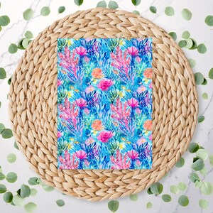 May include: A rectangular fabric with a vibrant watercolour design of an underwater coral reef. The pattern features pink, blue, and orange coral, set against a light blue background. The fabric is placed on a woven, circular placemat.