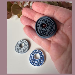 May include: Three decorative buttons with wave patterns. One large, dark gray button with a central hole and a smaller, red center is held in a hand. Two smaller, oval buttons, one gray and one blue, are also visible.