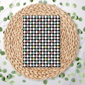 May include: A patterned notebook with a black and white checkered design, adorned with colorful hearts, diamonds, and floral shapes. The notebook rests on a woven placemat, surrounded by green leaves.