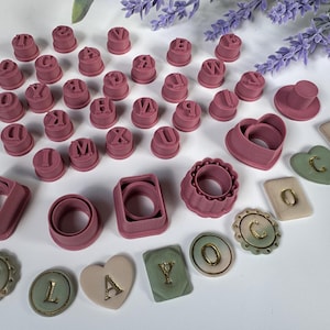 May include: A collection of pink plastic alphabet letter stamps, various geometric cookie cutters, and finished clay pieces in heart, circle, and rectangular shapes. The finished pieces feature gold-coloured letters spelling CLAYOCO.