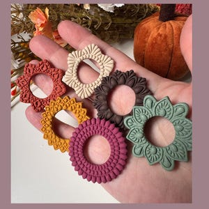 May include: A hand holds seven decorative, circular elements in various colours. The elements are in shades of red, yellow, brown, beige, and green. Each has a unique, detailed design with a central hole. A pumpkin and foliage are in the background.