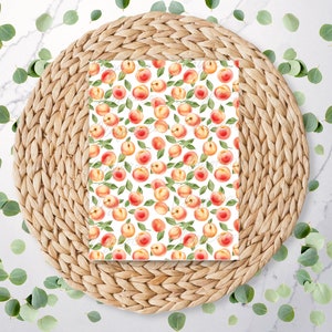 May include: A rectangular paper with a watercolor peach pattern, featuring red and yellow peaches with green leaves, rests on a woven, circular placemat. Eucalyptus leaves are scattered around the placemat on a white marble surface.