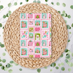 May include: A notebook with a repeating pattern of Christmas-themed postage stamps. The stamps feature festive elements such as snow globes, gingerbread houses, and mugs of cocoa, in pink, green, and teal. The notebook is on a woven, circular placemat.
