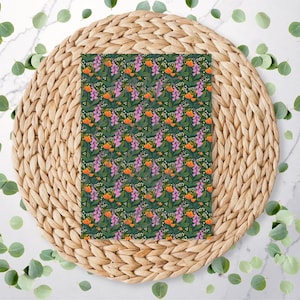 May include: A patterned fabric with a dark green background, featuring a repeating floral design of orange and purple flowers with white accents. The fabric is displayed on a woven, circular placemat, with scattered green leaves.