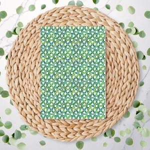 May include: A rectangular patterned paper with a green and white geometric design, resting on a woven, circular placemat. Green eucalyptus leaves are scattered around the placemat on a white marble surface.