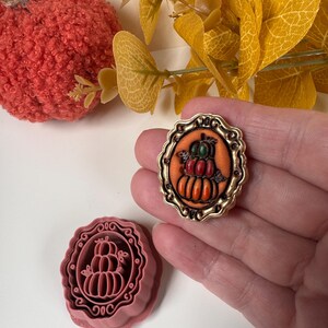 May include: A pink plastic cookie cutter featuring a stacked pumpkin design within an ornate scalloped frame, accompanied by a finished orange pumpkin-themed brooch with a metallic gold border and a detailed three-tiered pumpkin illustration.