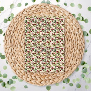 May include: A notebook with a cherry and leaf pattern, placed on a woven placemat. The notebook has a cream background with red cherries and green leaves. Eucalyptus leaves are scattered around the placemat.