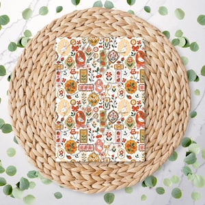 May include: A patterned paper sheet featuring a repeating design of swans, flowers, and decorative frames in red, orange, and cream colors. The paper is set on a woven, circular placemat with green leaves scattered around.