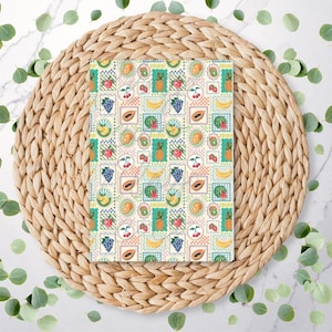 May include: A notebook featuring a vibrant fruit stamp design with illustrations of bananas, grapes, and cherries. It rests on a round, woven wicker placemat, with green leaves scattered around it.