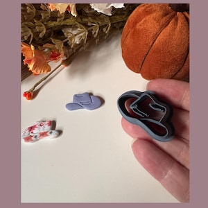 May include: A collection of miniature cowboy hat-shaped cookie cutters in various stages of completion. One is held in a hand, another is a finished product, and a third is a partially decorated design. The color palette includes shades of gray, lavender, and red.