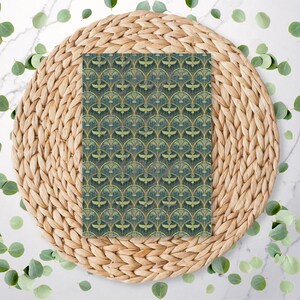 May include: A rectangular piece of paper featuring a repeating Art Deco design in green and gold hues. It rests on a circular woven placemat, with scattered green leaves on a white marble surface. The design is symmetrical.