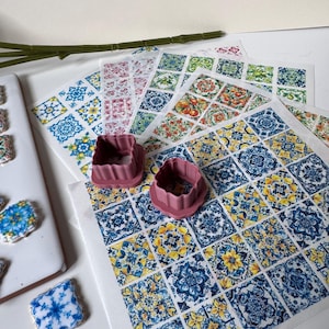 Peut inclure: Deux emporte-pièces carrés mauve à bords festonnés posés sur plusieurs feuilles de papier comestible présentant une grille de motifs de carreaux méditerranéens bleus, jaunes et orange, avec des biscuits carrés finis à proximité.