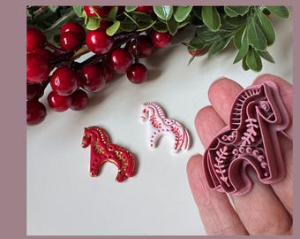 May include: Three decorative horse figurines in red, white, and pink with gold accents. One is a cookie cutter held in a hand. The background includes red berries and green leaves.