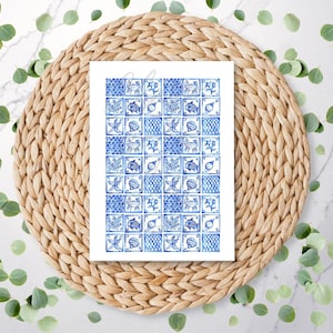 May include: A rectangular print with a blue and white tile pattern, depicting marine life. It rests on a circular woven mat, with green eucalyptus leaves scattered around on a white marble surface.
