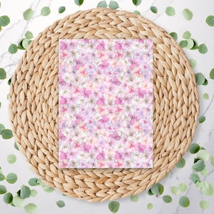 Peut inclure: Un motif floral rose et blanc avec un effet aquarelle. Le motif est sur un morceau de papier carré et est posé sur un tapis de paille tissé.