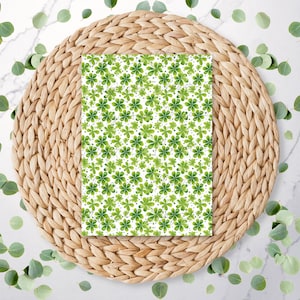 May include: A rectangular white card with a repeating pattern of green four-leaf clovers. The card is centered on a round, woven, light brown placemat. Green eucalyptus leaves are scattered around the placemat on a white marble surface.