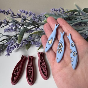 May include: A selection of fish-shaped craft items. Three pale blue fish pendants with gold detailing are held in a hand. Three maroon fish-shaped cookie cutters are also visible. The background includes lavender sprigs.