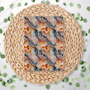 May include: A rectangular item with a repeating pattern of agate-like designs in shades of orange, blue, and gold. The item is centered on a round, woven placemat. Green leaves are scattered around the placemat on a white marble surface.