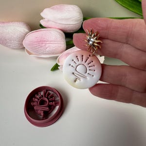 May include: A gold earring with a sunburst design and a clear gemstone, alongside a white and yellow pendant featuring a sun and rays design. A matching pink cookie cutter is also visible, with pink tulips in the background.
