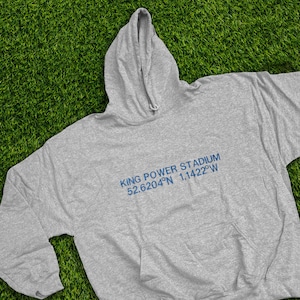 Puede incluir: Sudadera con capucha gris con bolsillo de canguro. El texto "KING POWER STADIUM 52.6204°N 1.1422°W" está impreso en azul. La sudadera tiene una capucha con cordón y está hecha de un material suave.