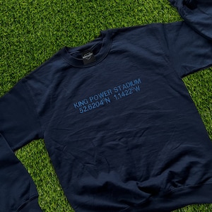 Puede incluir: Sudadera azul marino con el texto "KING POWER STADIUM 52.6204°N 1.1422°W" en azul claro. La sudadera tiene cuello redondo y mangas largas. La marca "MATCHDAY" está en la etiqueta del cuello. La sudadera está sobre una superficie verde.