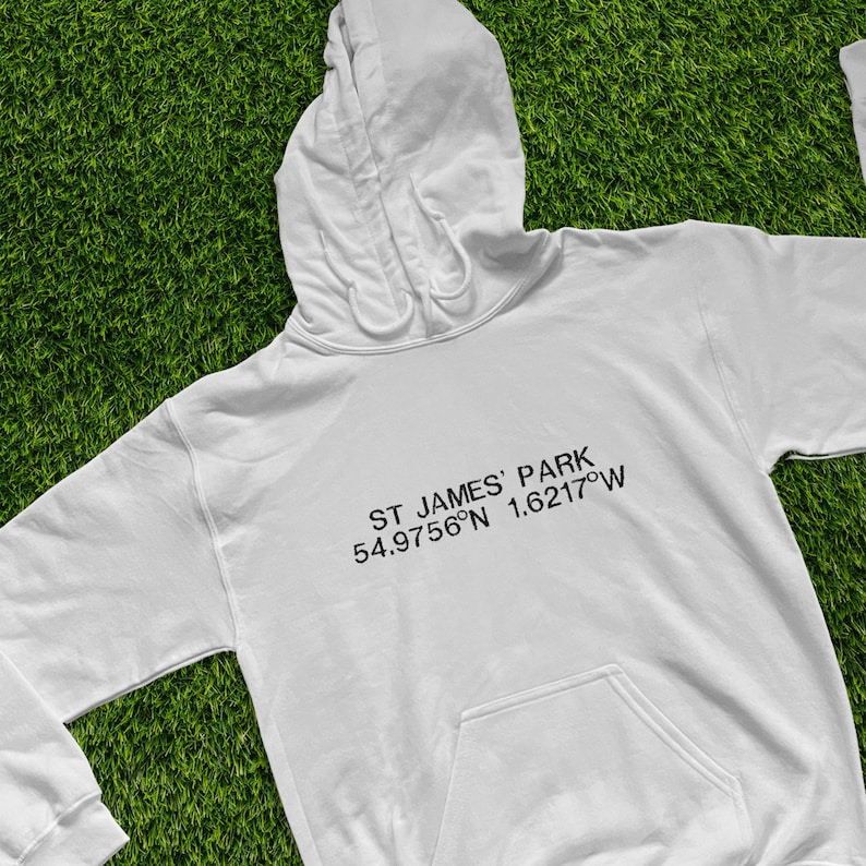 May include: White hooded sweatshirt with a front pocket and drawstring hood. The text "ST JAMES' PARK 54.9756&deg;N 1.6217&deg;W" is printed in black on the front. The sweatshirt is displayed on a green grassy surface.