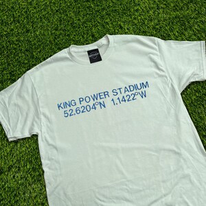 Puede incluir: Camiseta blanca con el texto azul "KING POWER STADIUM 52.6204°N 1.1422°W". La camiseta tiene cuello redondo y mangas cortas. Una pequeña etiqueta negra con texto blanco es visible en el cuello.