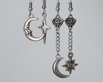 May include: A set of three pairs of silver earrings. The first pair features a crescent moon with a face. The second pair features a star. The third pair features a dangling eye charm with a crescent moon and star charm at the bottom.