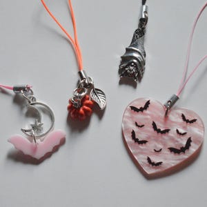 May include: A collection of Halloween-themed charms. Includes a pink bat charm with a crescent moon and star, a red pumpkin charm with a leaf, a silver bat charm, and a pink heart charm with black bat silhouettes.