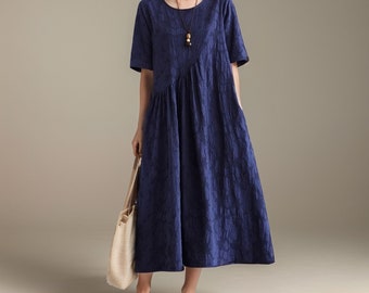 Puede incluir: Un vestido midi azul marino con un patrón texturizado y un dobladillo ligeramente asimétrico. El vestido tiene mangas cortas y un escote redondo. La modelo lleva el vestido con un collar marrón y un bolso de mano beige.